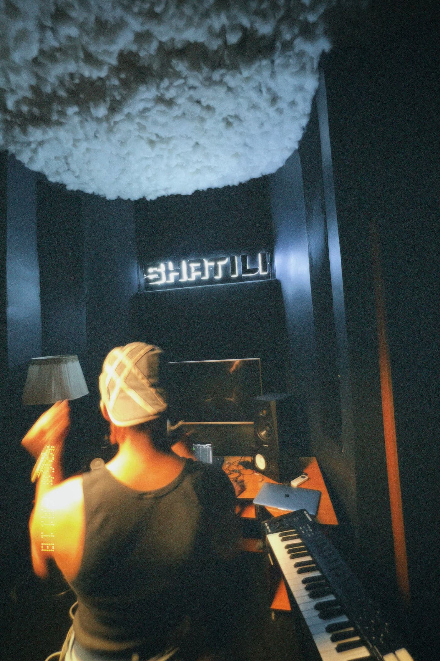An artist at the keyboard in a dark studio beneath a lit SHATILI sign.